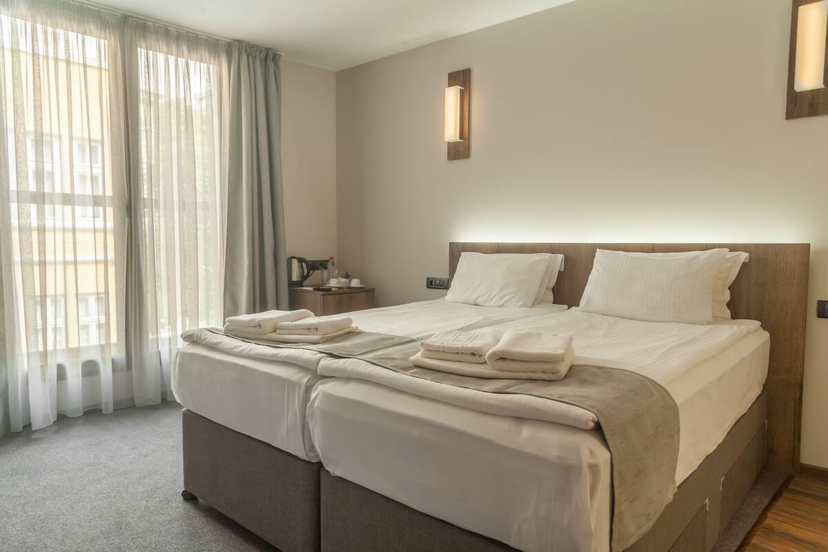 Quarto de hotel com uma cama grande, mesa de cabeceira com abajur e cortinas finas na janela.