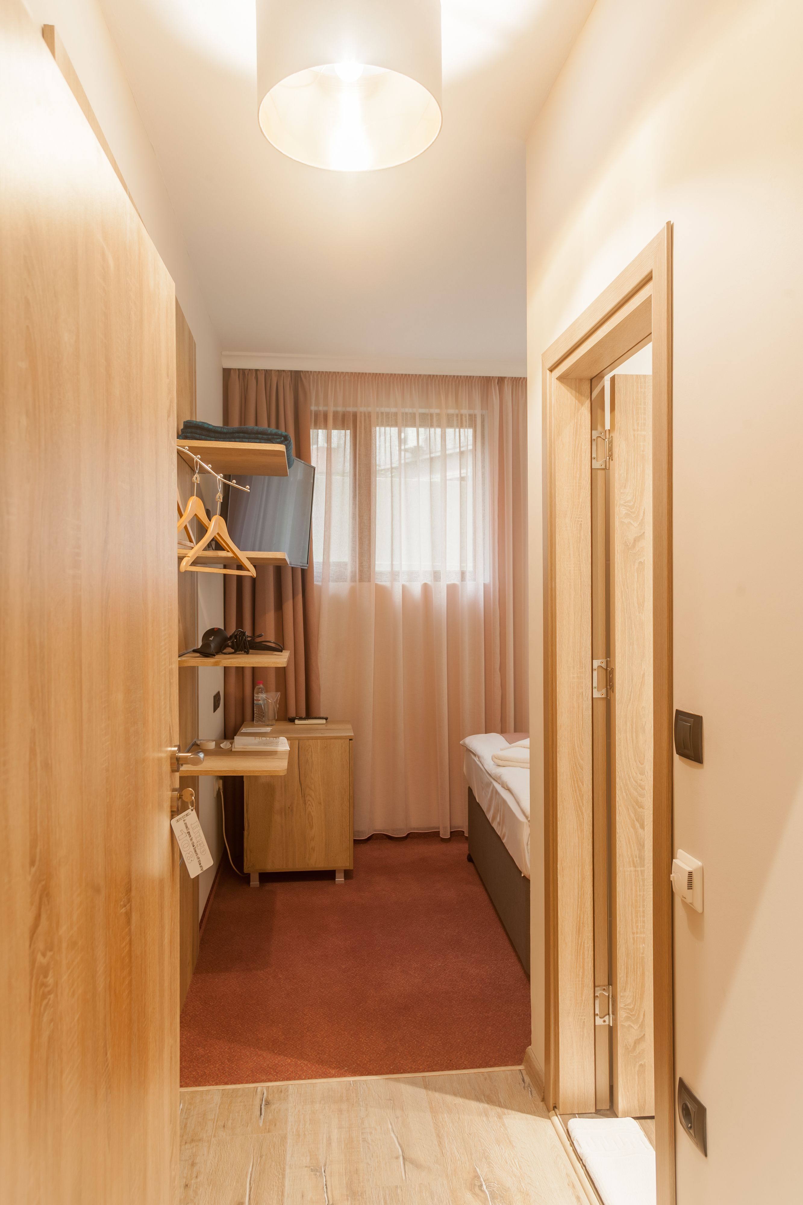 A compact hotel room with a bed, a small desk, a wardrobe, and a hanging clothes rack.