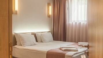 Quarto de hotel com cama, piso de madeira, cortinas e luminárias fixadas na parede.