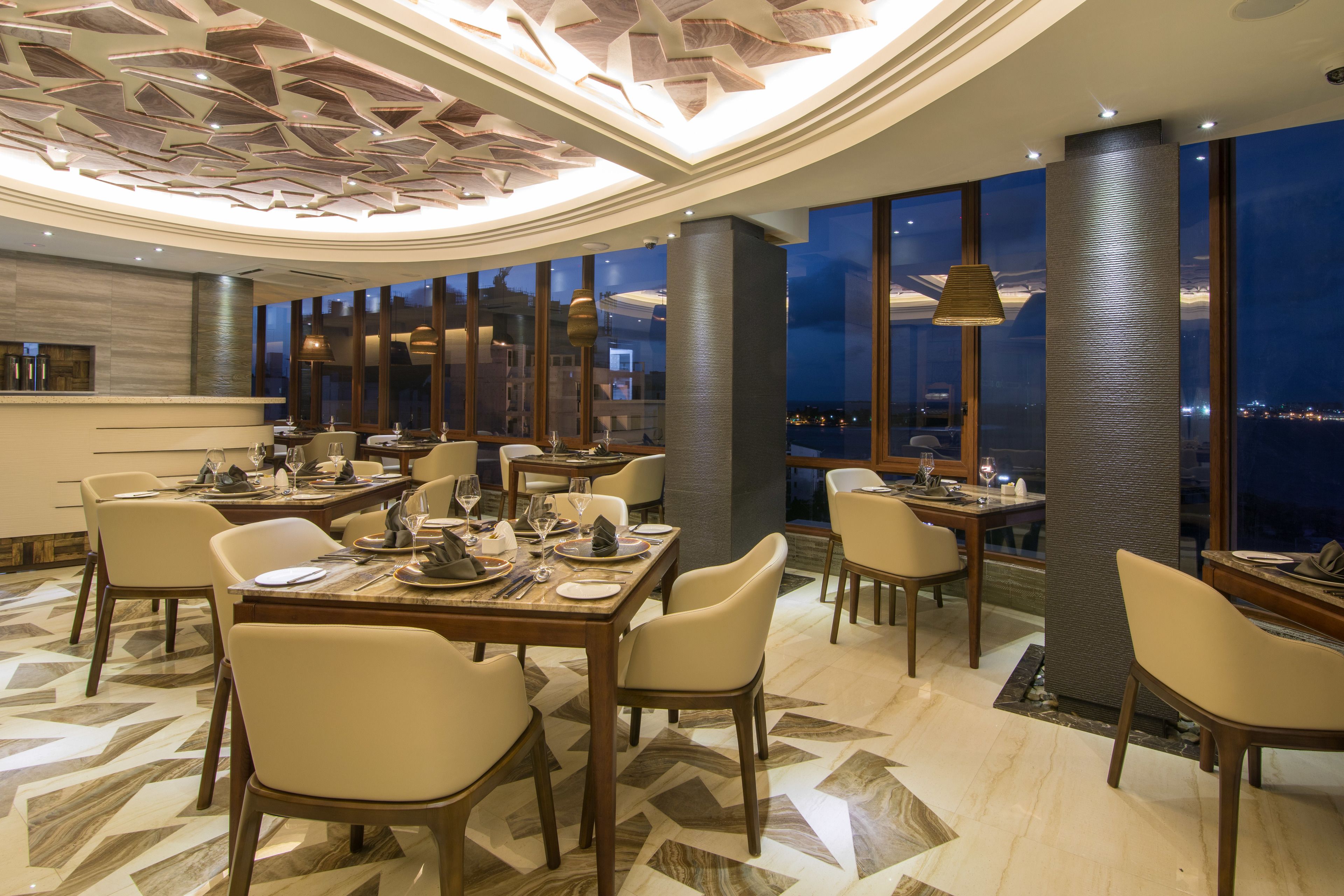 A modern dining area with multiple tables and chairs, a patterned floor, and large windows offering a city view.