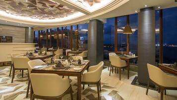 A modern dining area with multiple tables and chairs, a patterned floor, and large windows offering a city view.