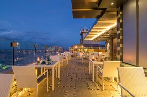Un área de comedor al aire libre con mesas y sillas, vista del paisaje urbano y un edificio moderno al fondo.
