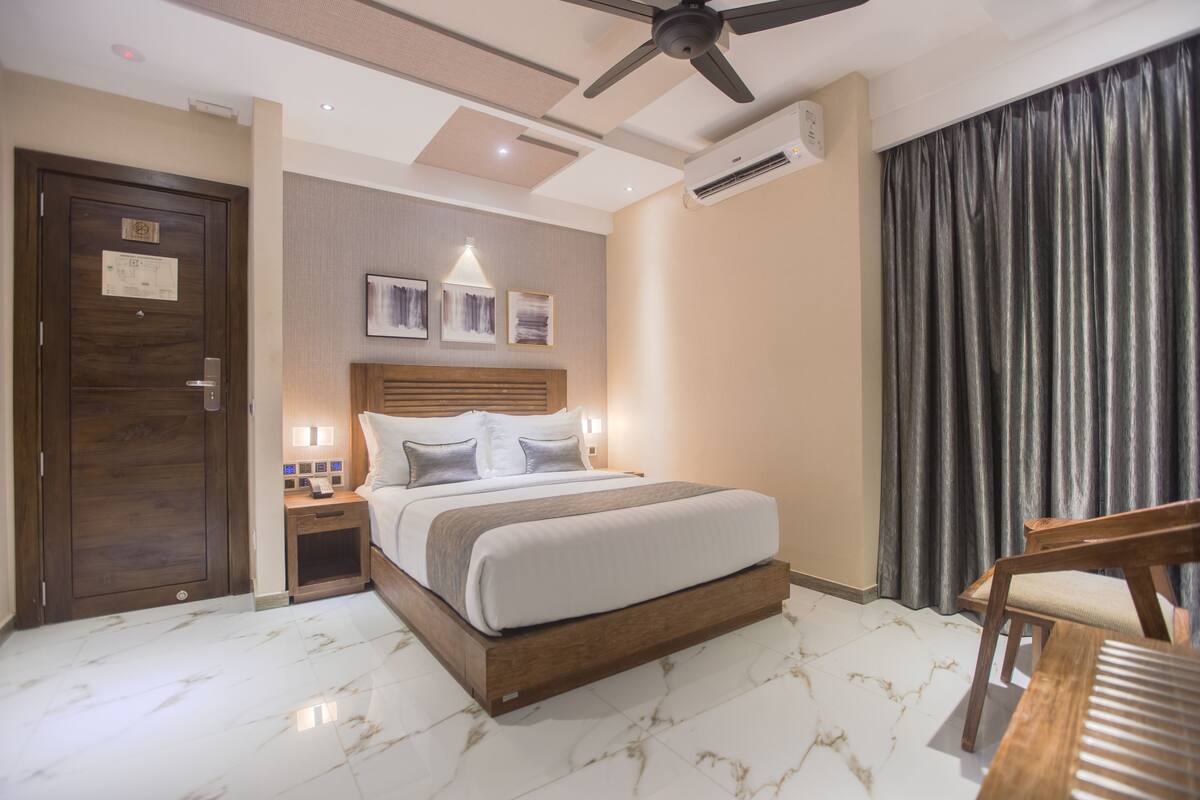 A hotel room with a large bed, a wooden nightstand, a desk, a ceiling fan, and a window with curtains.