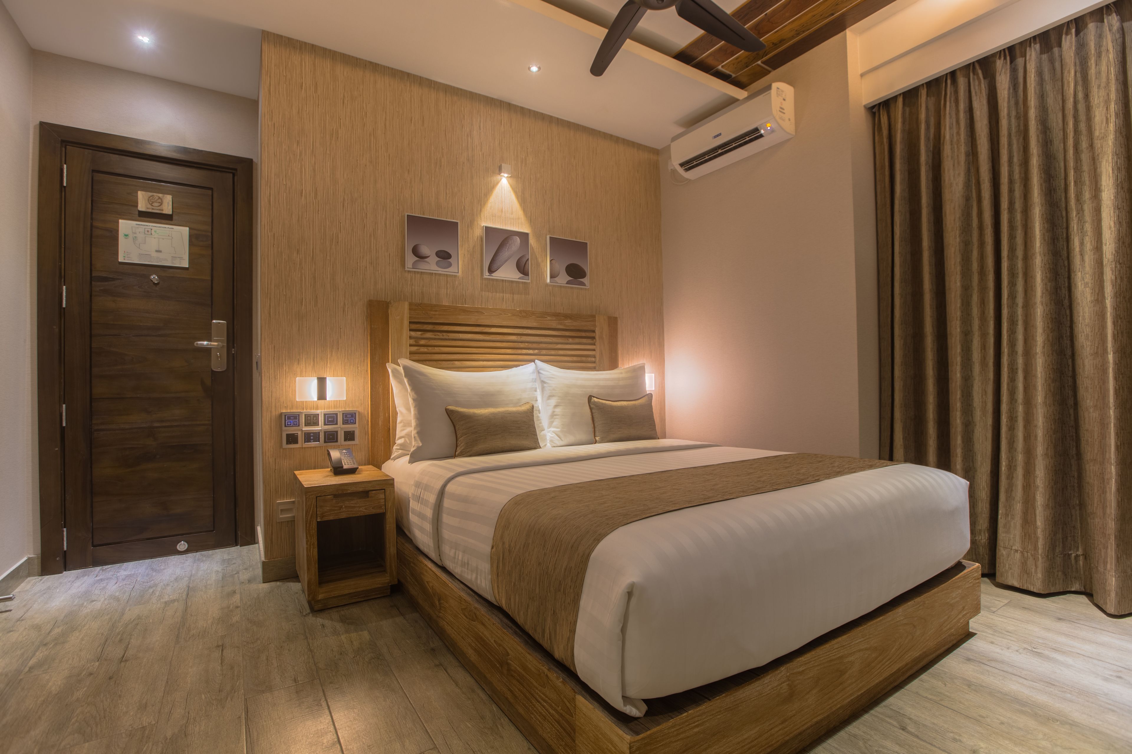 A modern hotel room with a wooden headboard, a large bed, a nightstand with a lamp, and a wooden floor.