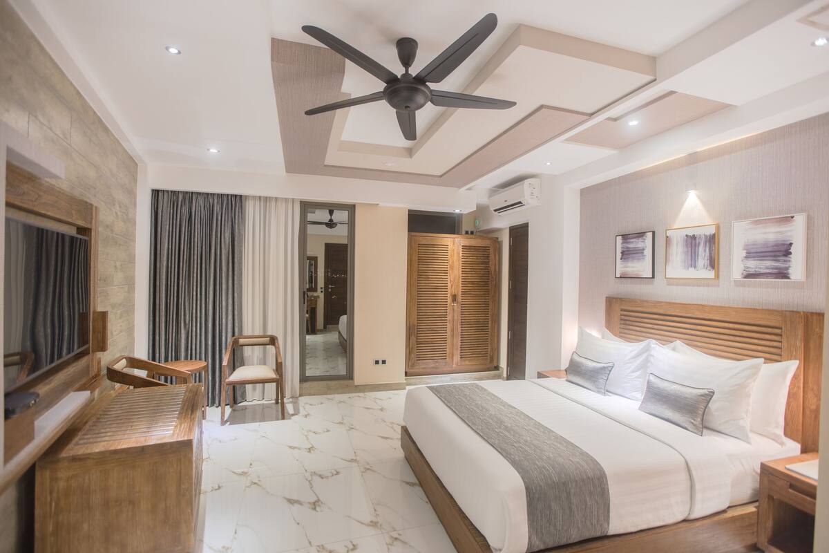 A modern hotel room with a large bed, a ceiling fan, a wooden desk, a chair, and a wardrobe.
