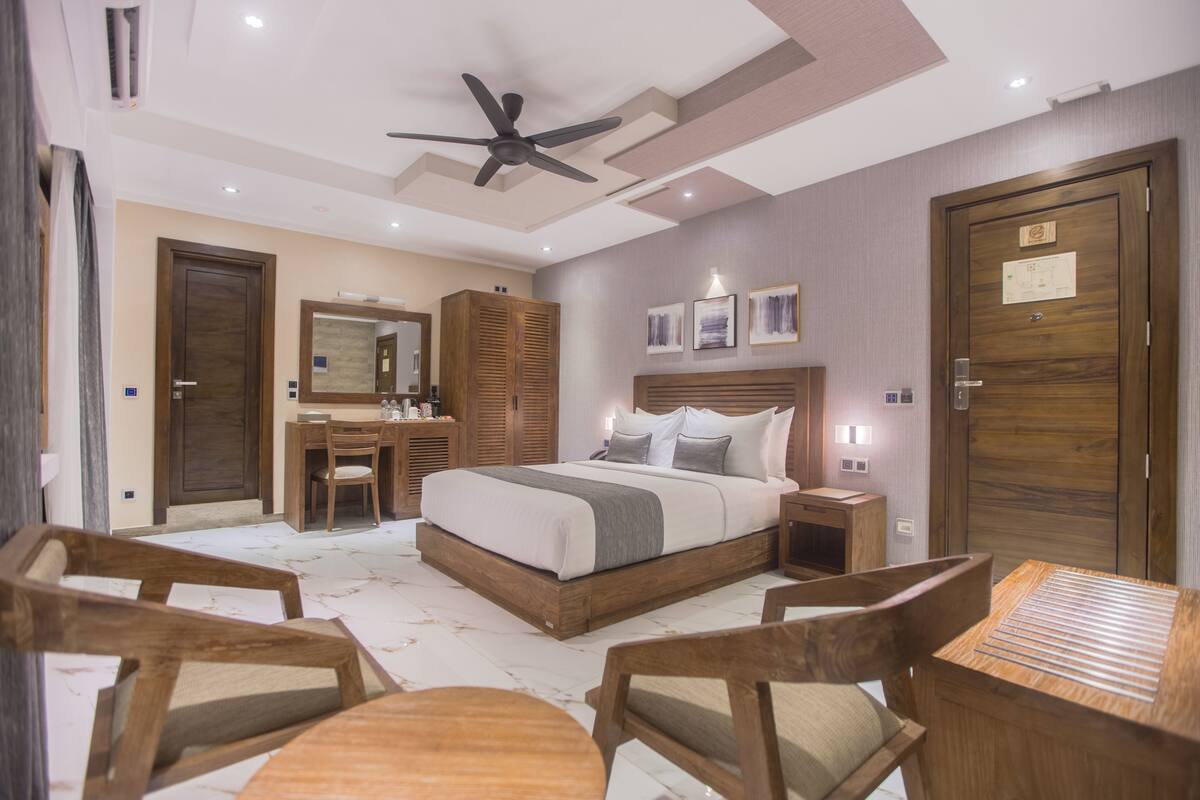 A modern hotel room with a large bed, wooden furniture, a ceiling fan, and a wooden door.