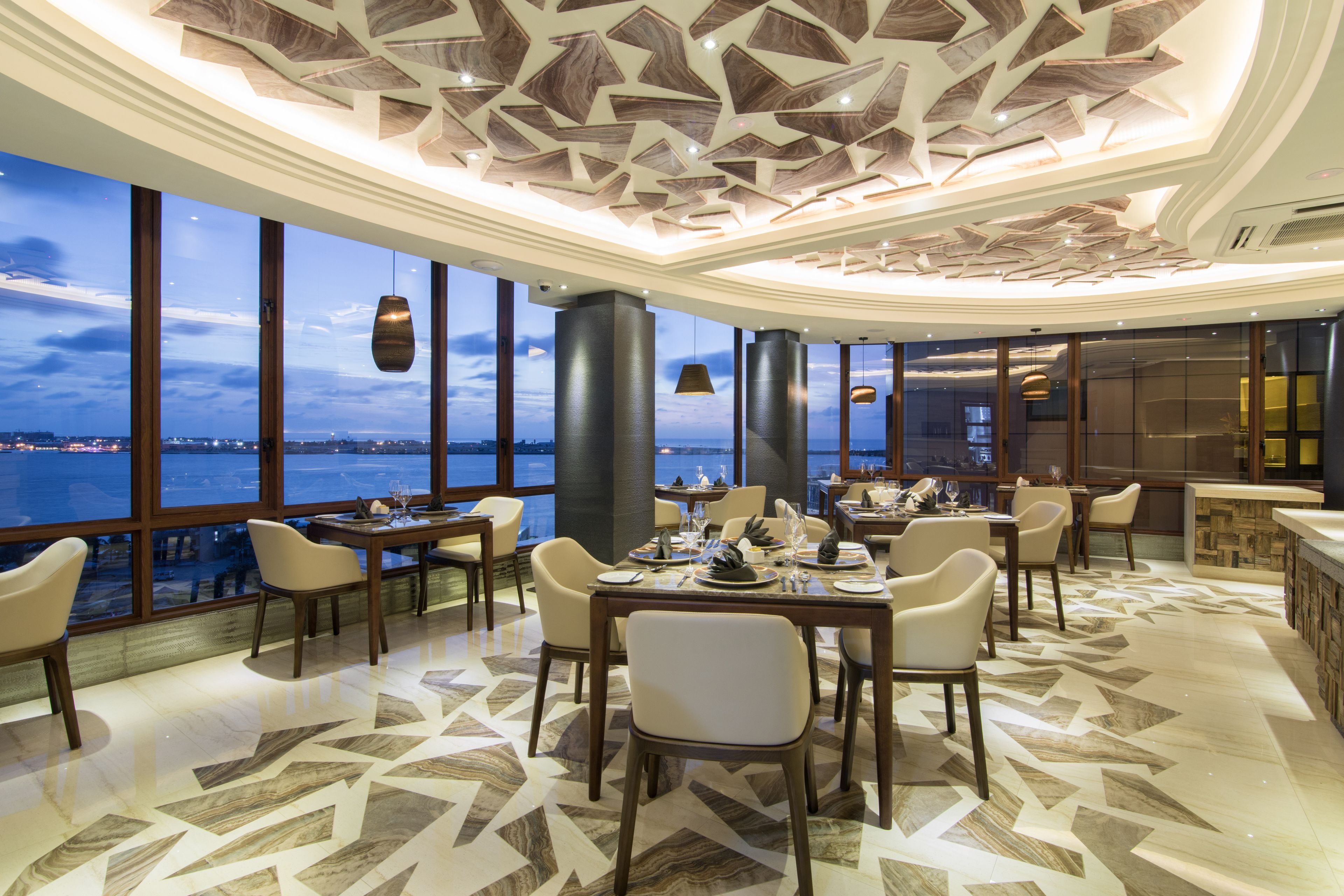 A modern dining area with a panoramic view of the water, featuring geometric ceiling design and contemporary furniture.