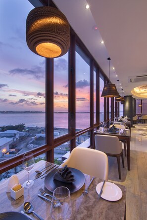 A restaurant with a view of the sea at sunset, featuring large windows, modern lighting, and a clean dining area. - Samann Grand (Malé)