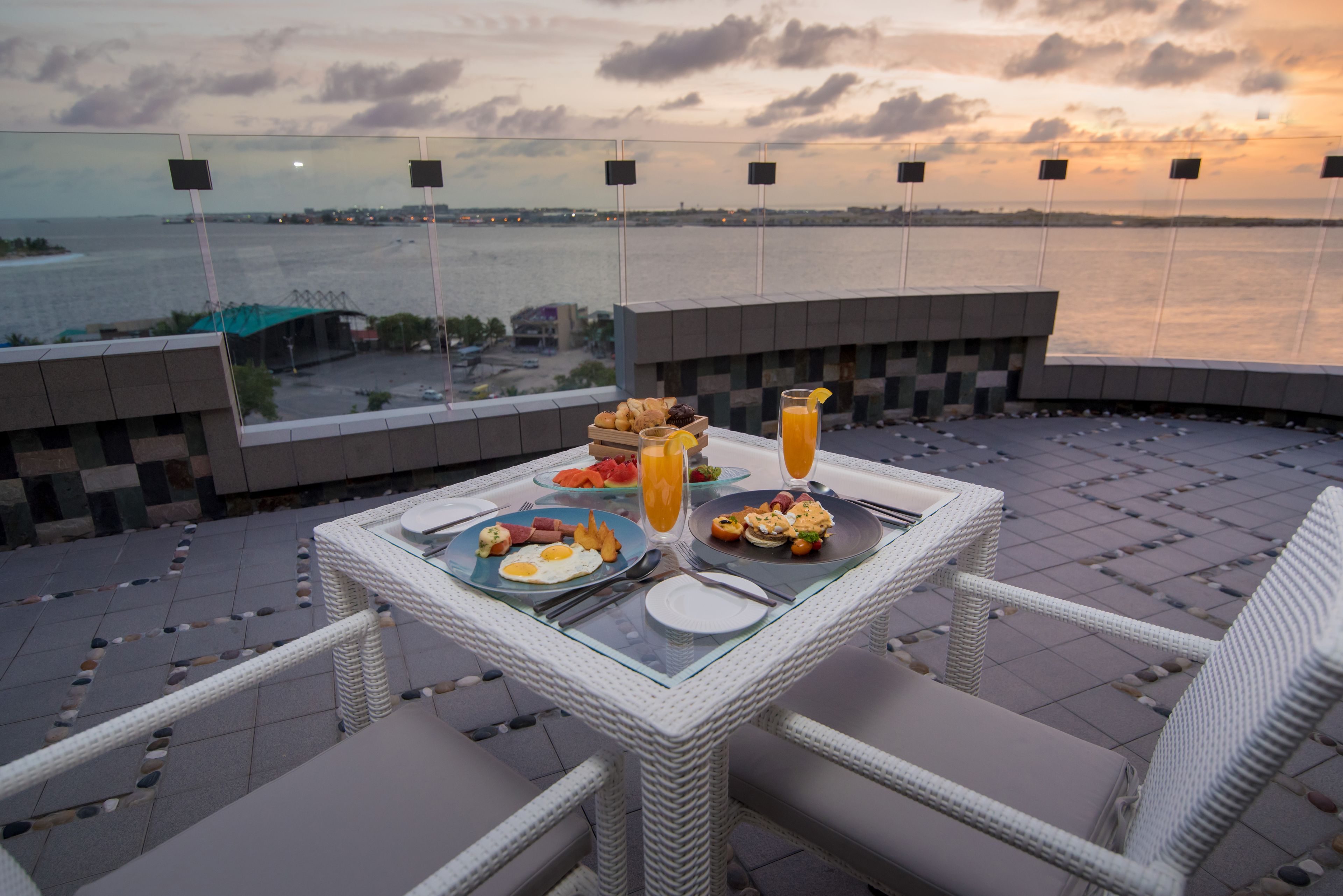 Mesa de comedor con alimentos para el desayuno y un vaso de jugo de naranja, con vista a un cuerpo de agua y un atardecer.