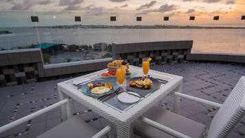 Mesa de comedor con alimentos para el desayuno y un vaso de jugo de naranja, con vista a un cuerpo de agua y un atardecer.