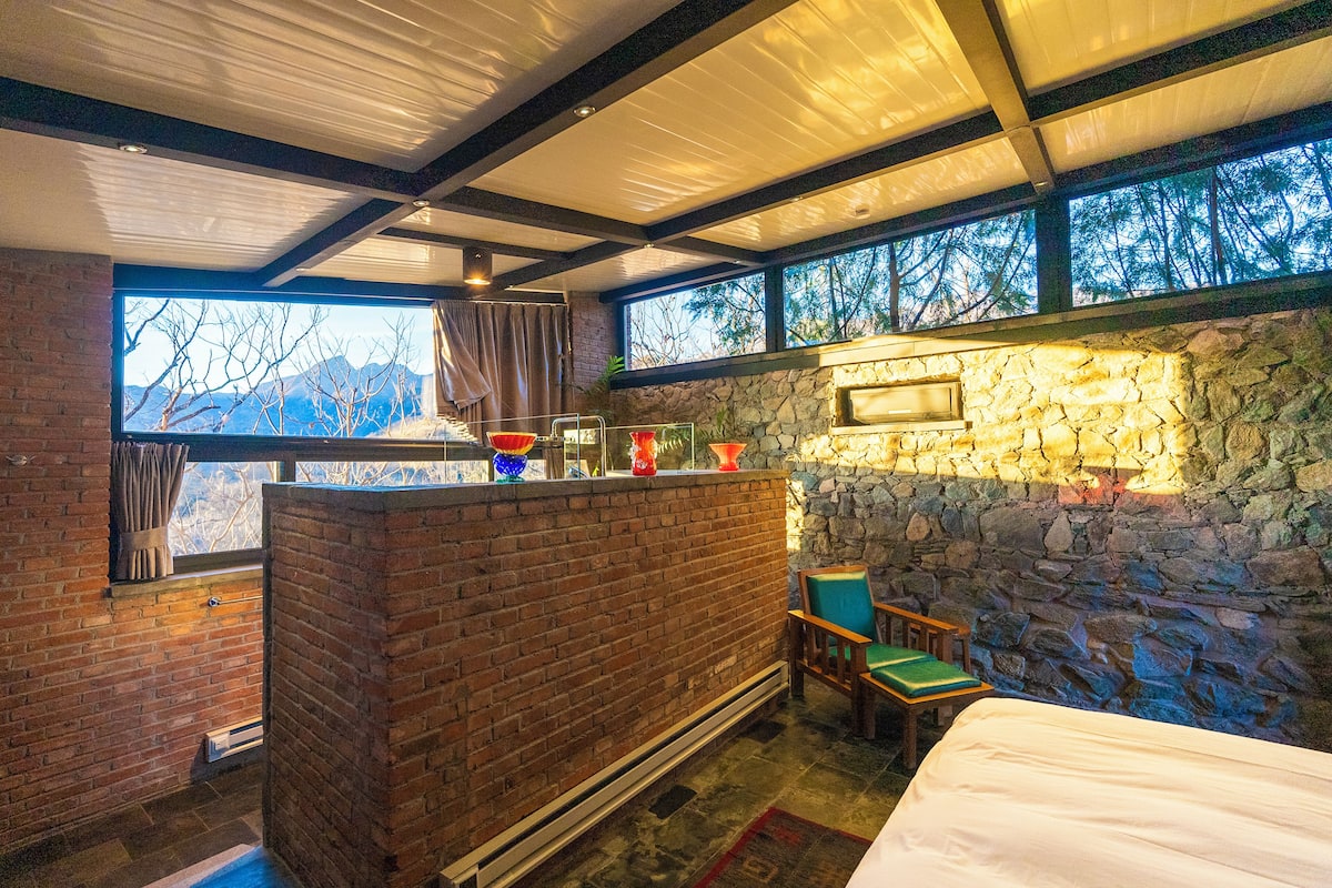A room with a stone wall, a brick counter, a bed, and a view of mountains through large windows.
