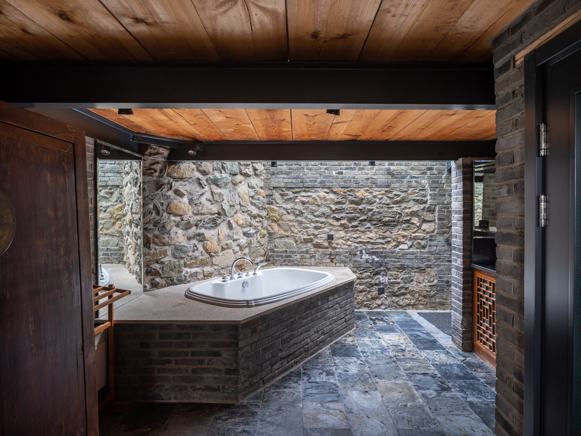 A modern bathroom with a stone wall, a wooden ceiling, and a large bathtub.