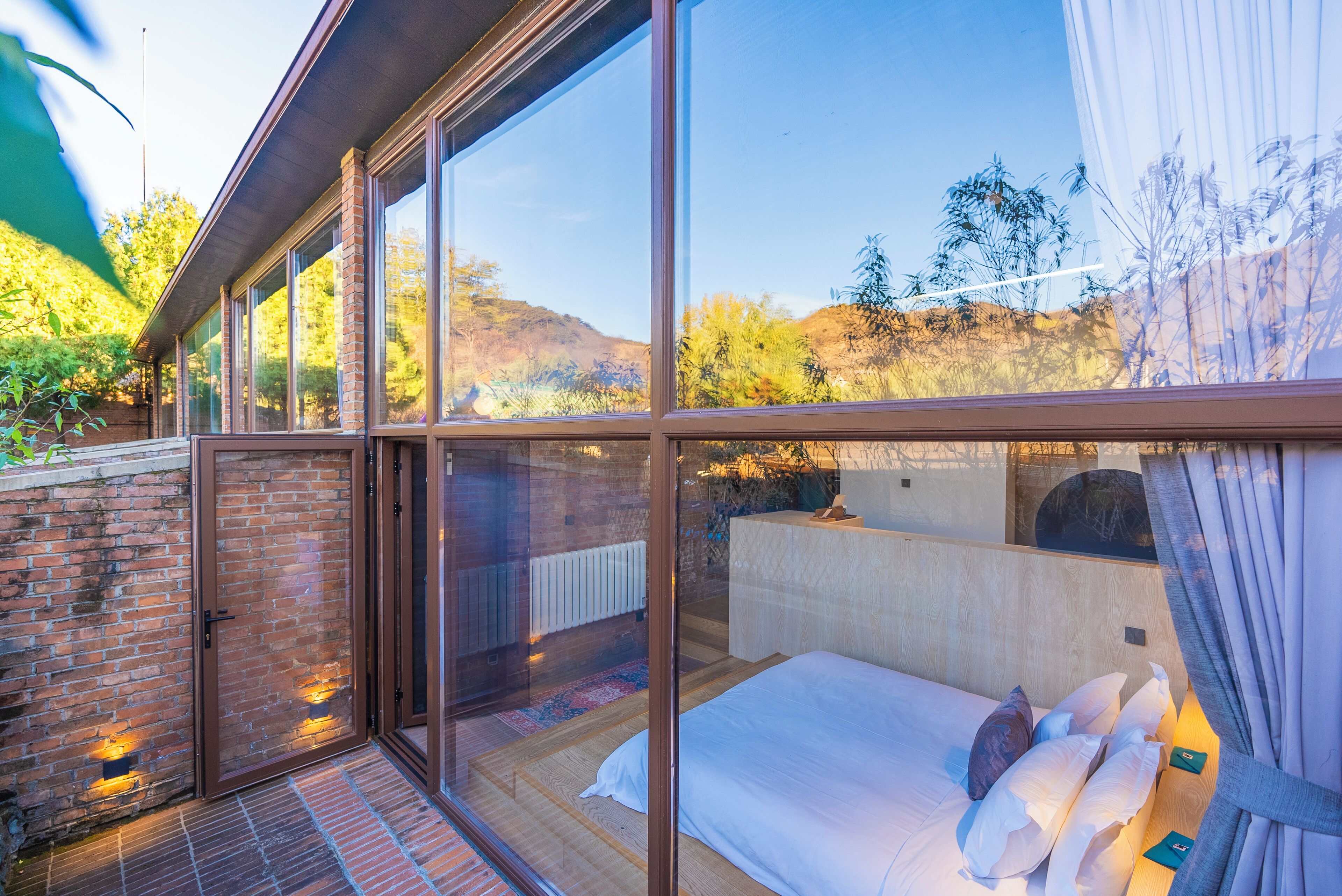 A modern bedroom with a large glass wall, a wooden floor, and a bed with pillows.