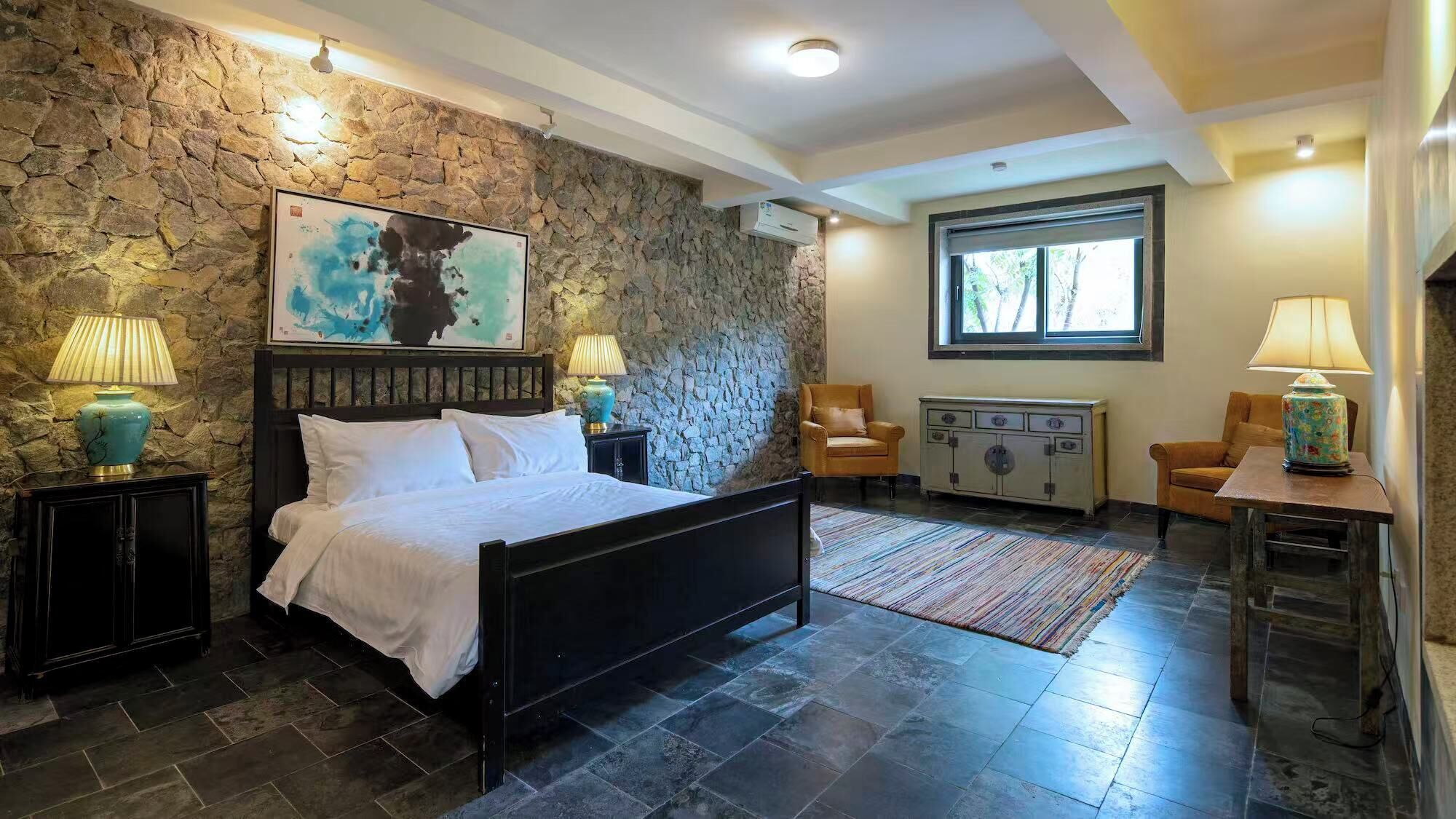 A bedroom with a stone wall, a bed, two chairs, a dresser, and a window.