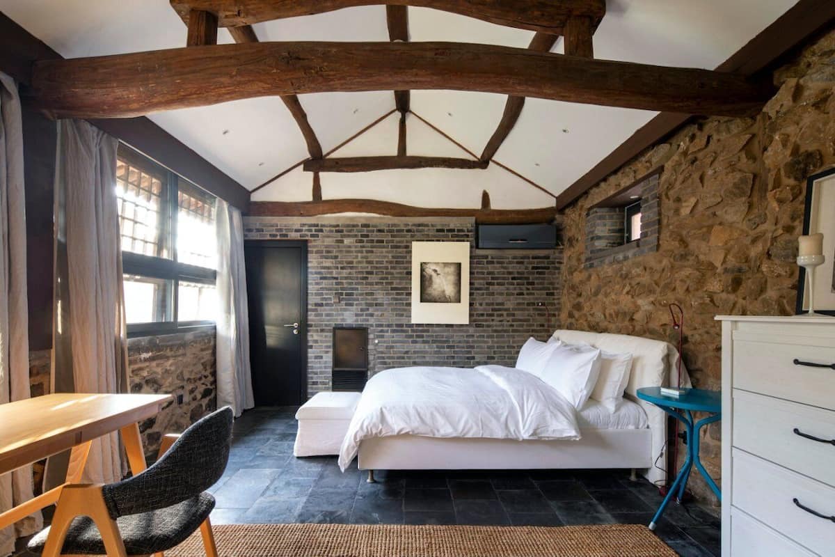 A bedroom with a large bed, a desk, and a chair. There is a stone wall and a wooden ceiling.