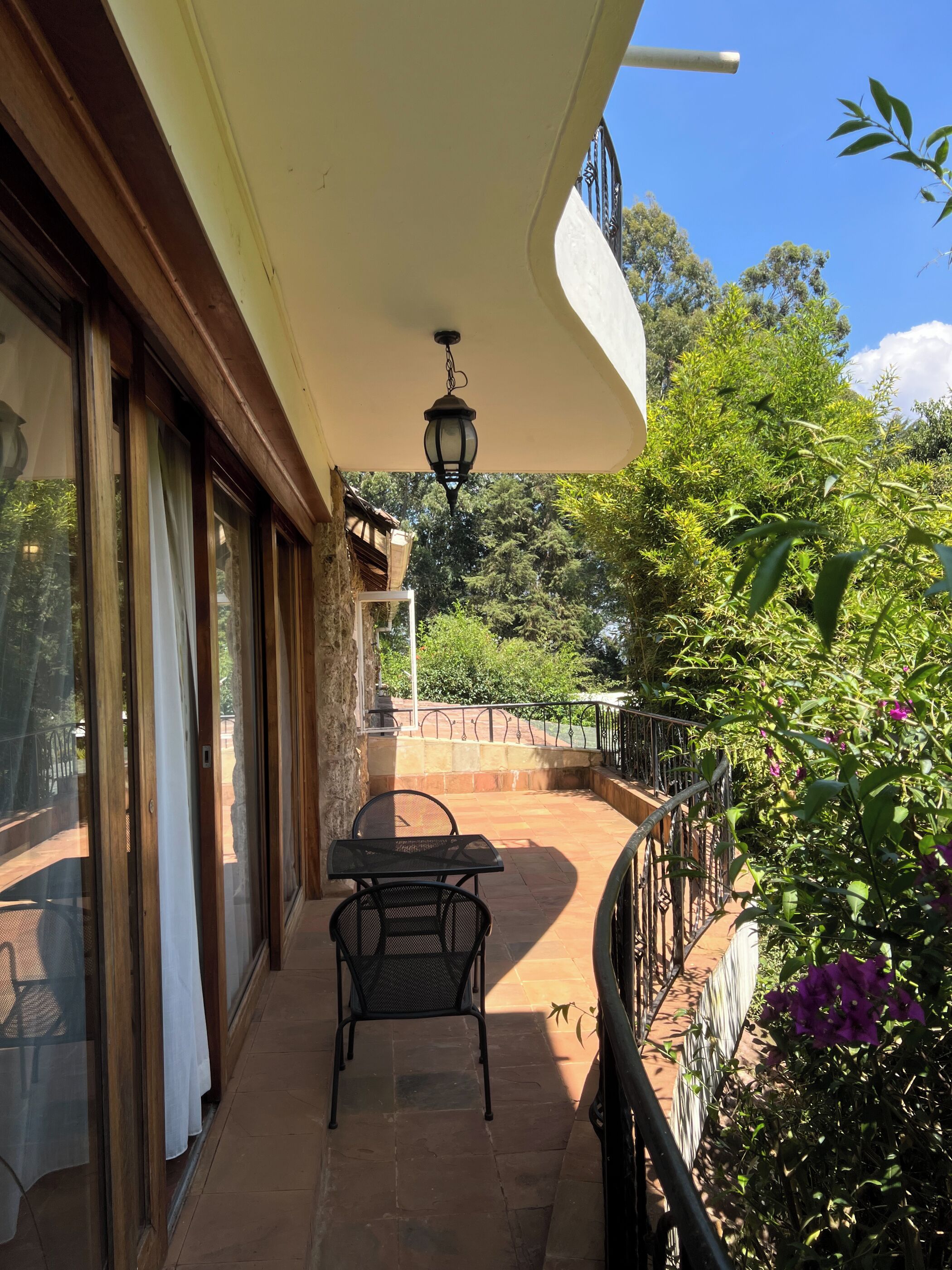 A covered outdoor patio with a table and chairs, a hanging light fixture, and a view of greenery and trees.