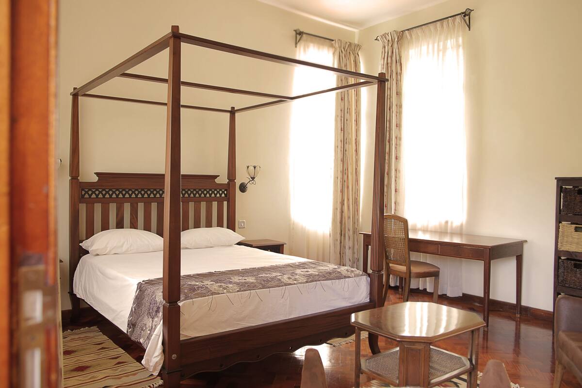 Uma cama de dossel com cabeceira de madeira, uma escrivaninha com cadeira e uma mesa de cabeceira.