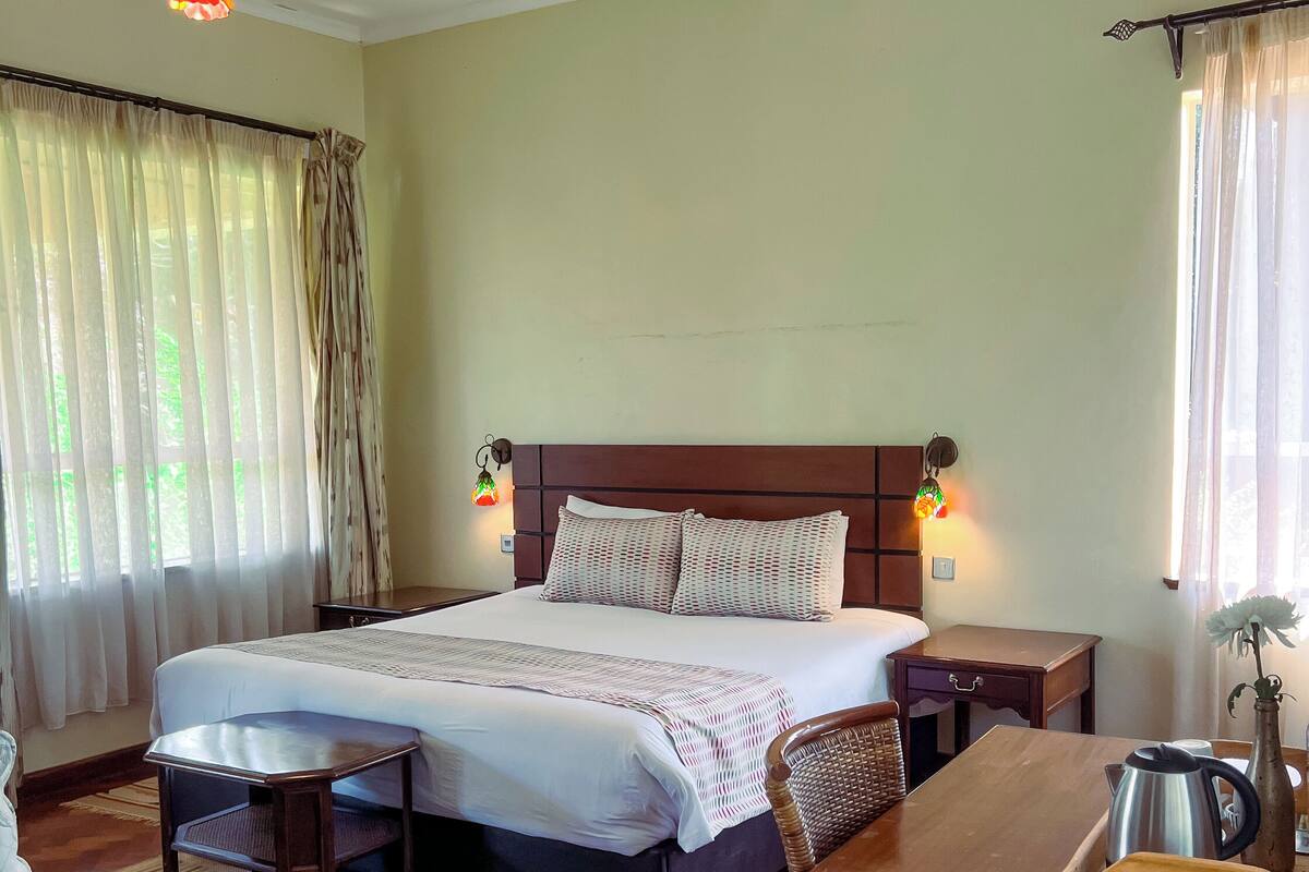 Quarto de hotel com uma cama grande, mesas de cabeceira, uma mesinha de madeira, escrivaninha, cadeira e um tapete estampado.