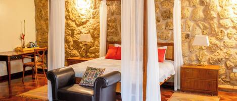 A bedroom with a four-poster bed, a leather armchair, a nightstand, and a stone wall.