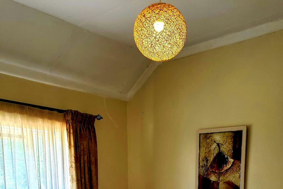 Quarto com cama grande, cabeceira de madeira, mesa de cabeceira, tapete estampado, janela com cortinas e luminária pendente.