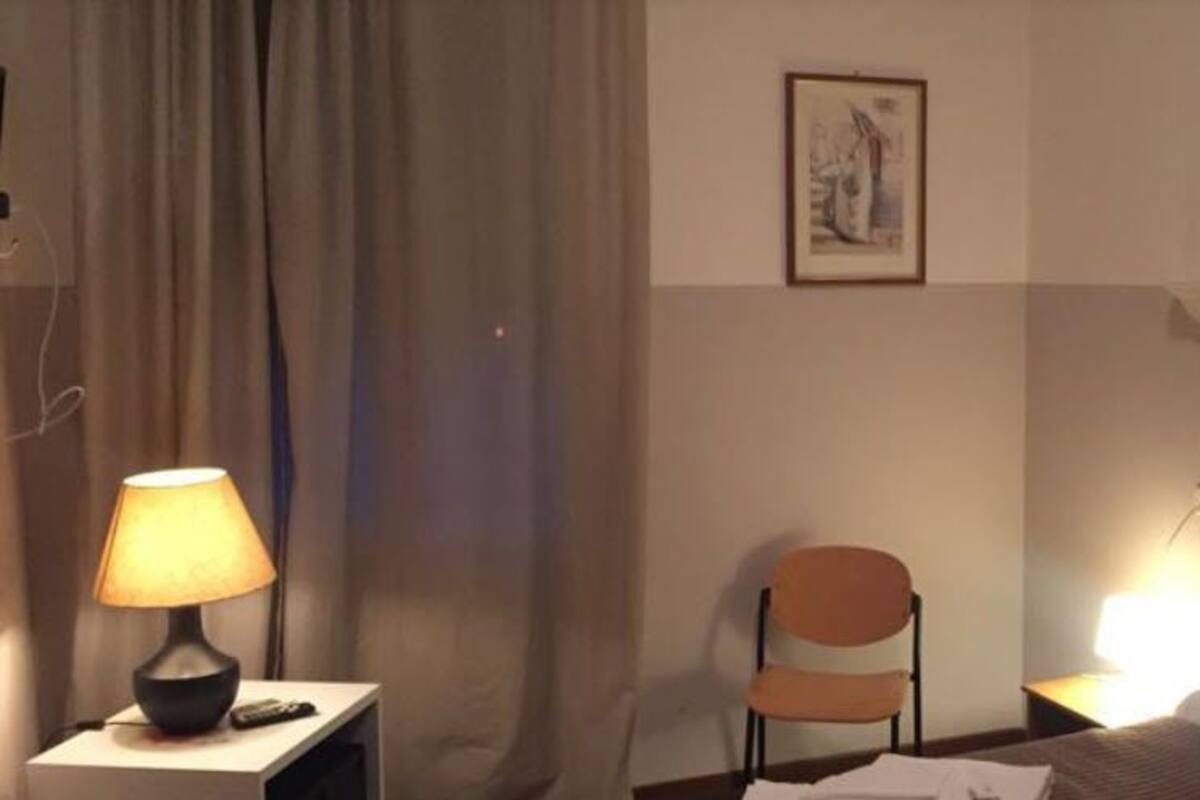 Hotellihuone, jossa on sänky, tuoli, lamppu, seinällä oleva taulu ja juliste.