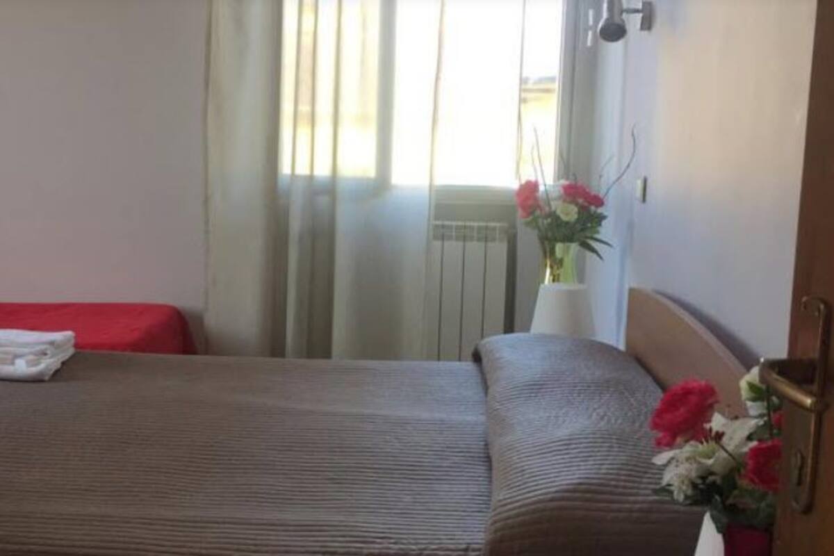 Hotellihuone, jossa on kaksi sänkyä, ikkuna verhoilla, patteri ja maljakko, jossa on kukkia.
