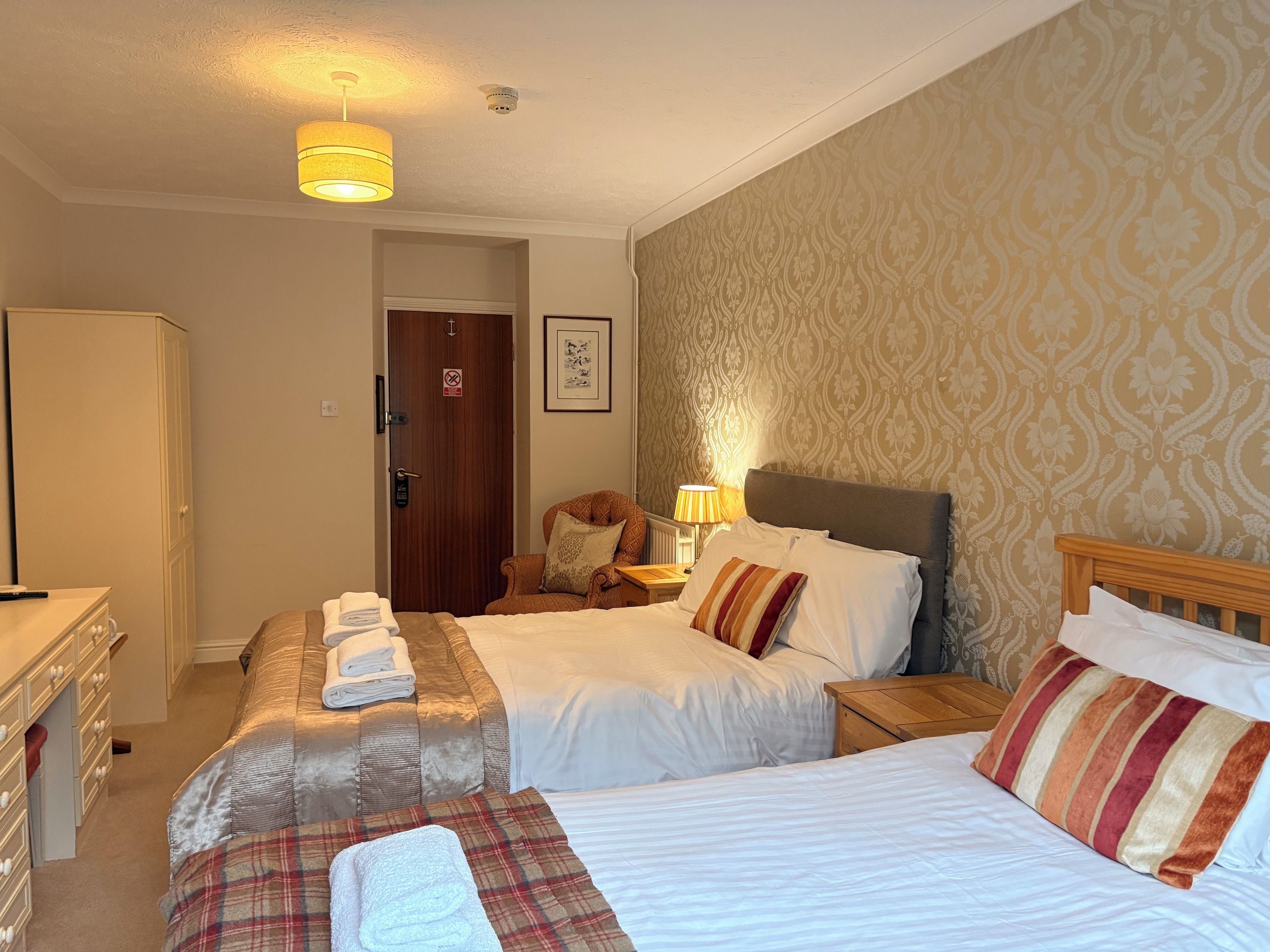 A hotel room with two beds, a wooden wardrobe, a desk with a chair, and a patterned wallpaper.