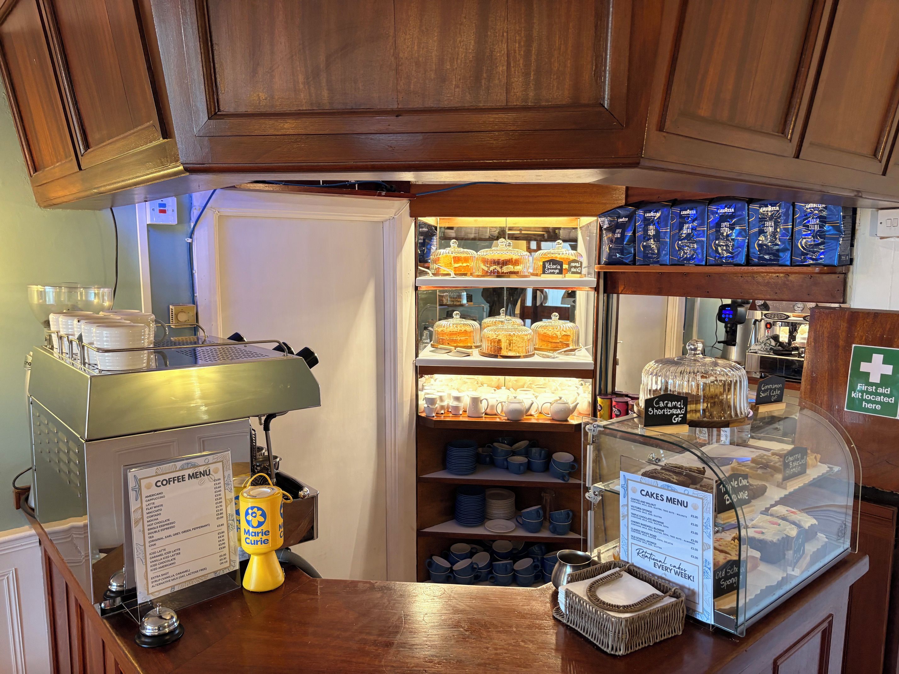 Bancada de café com cardápio, máquina de café e diversos produtos de panificação expostos em vitrine.