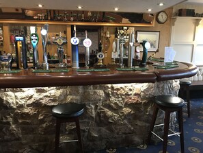 Zona de bar con varias griferías de cerveza, taburetes y un fondo de pared de piedra.