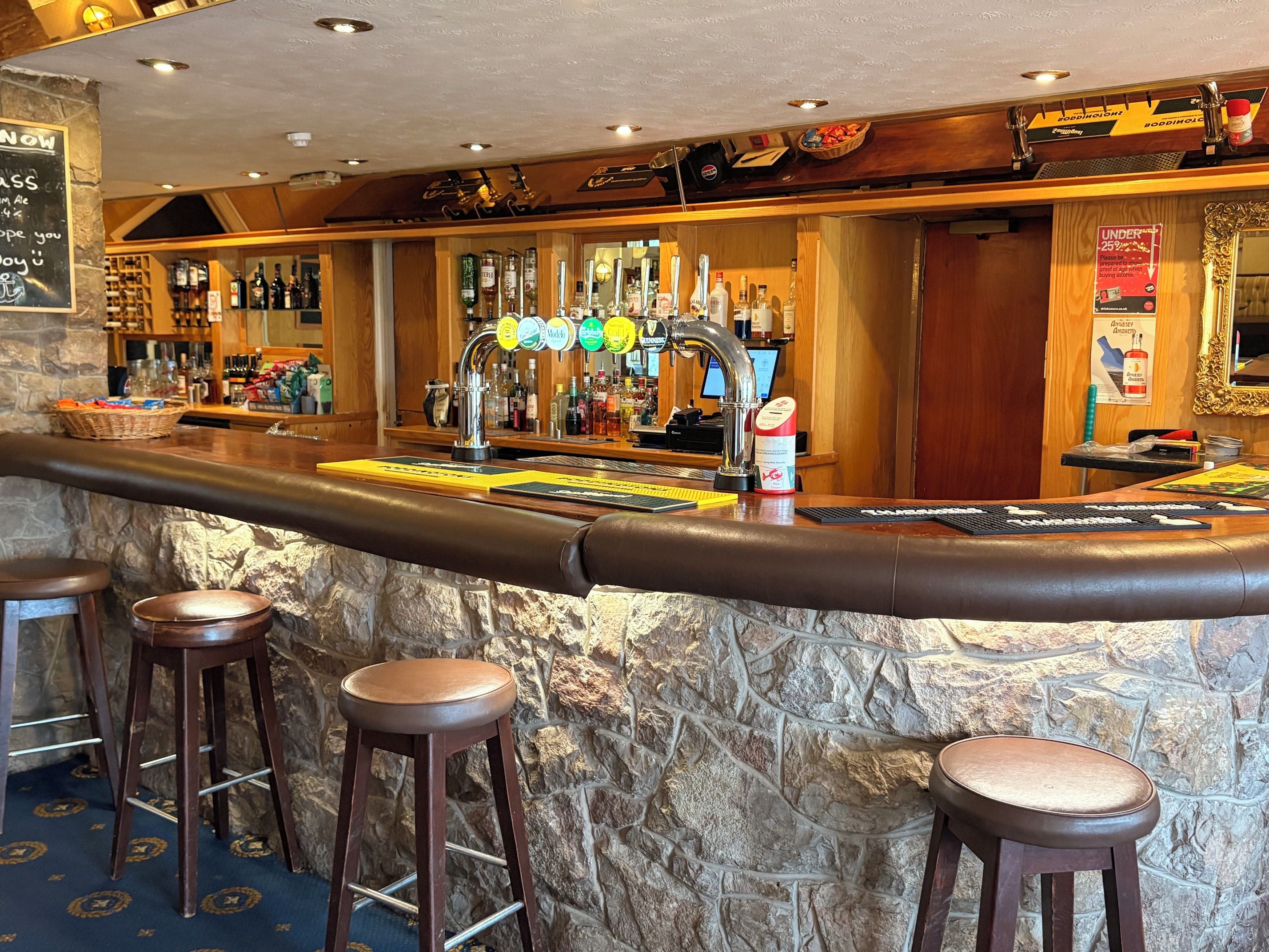 A bar area with a stone wall, stools, a bar counter, and a variety of bottles and glasses.