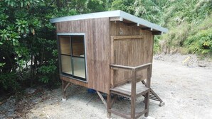 A wooden cabin with a large window, a small porch, and a dirt ground.