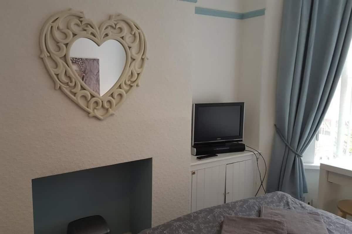 Um quarto com um espelho em formato de coração, uma televisão sobre um móvel, uma cama com manta e uma janela com cortinas.