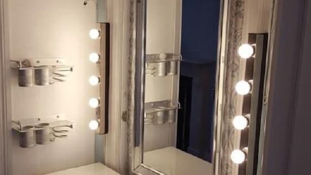 Une coiffeuse dotée d’un grand miroir, entouré de lumières, et d’une étagère avec des récipients.