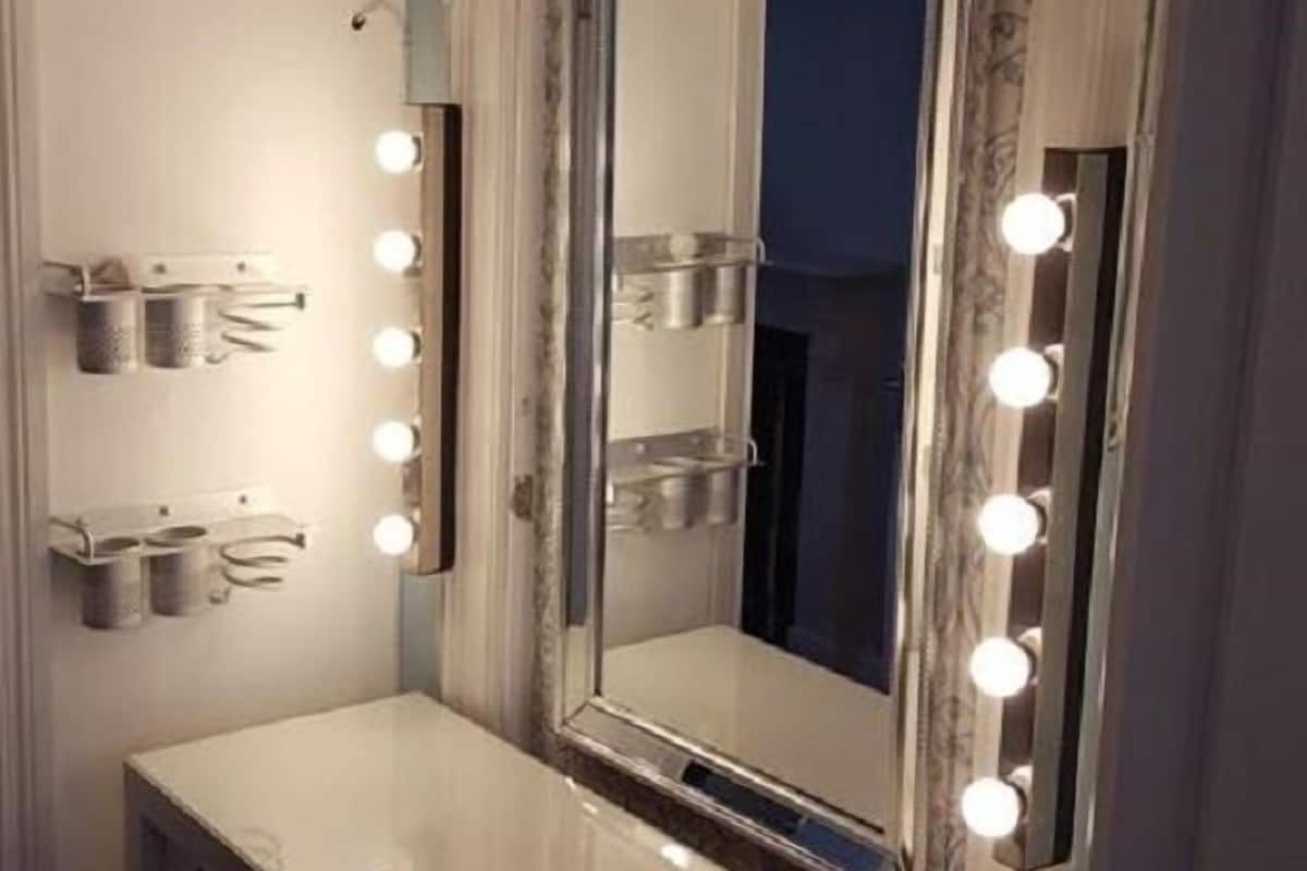 Uma toucador com um espelho grande, rodeado por luzes, e uma prateleira com recipientes.