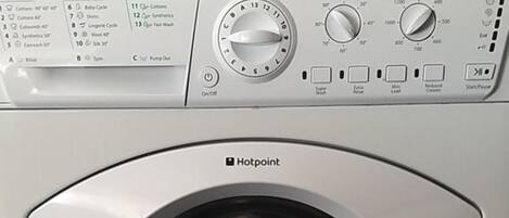 Vista frontal de una lavadora Hotpoint, con panel de control, toma de agua y manguera de desagüe.