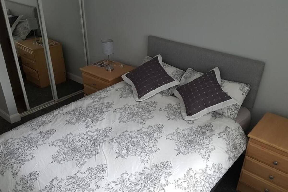 Ein ordentlich bezogenes Bett mit gemusterten Betttextilien, ein Nachttisch mit Lampe und ein Spiegel an der Wand.