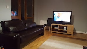 A living room with a dark leather sofa, a wooden entertainment unit with a television, and a radiator. - Alexandra Park Guest House (Belfast)