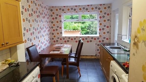 A compact kitchen with a dining table, chairs, a washing machine, and a window with a view of greenery. - Alexandra Park Guest House (Belfast)