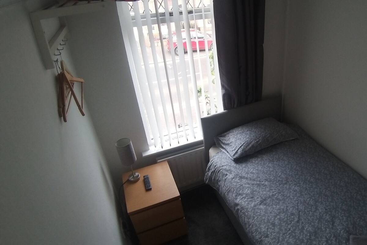 Ein kleiner, schlicht eingerichteter Raum mit einem Bett, einem Nachttisch, einer Lampe und einem Fenster mit Gitter.