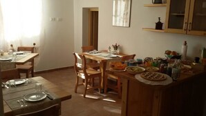 Zona de menjador amb taules i cadires de fusta, un taulell de cuina amb diversos aliments, i una finestra amb cortines transparents.