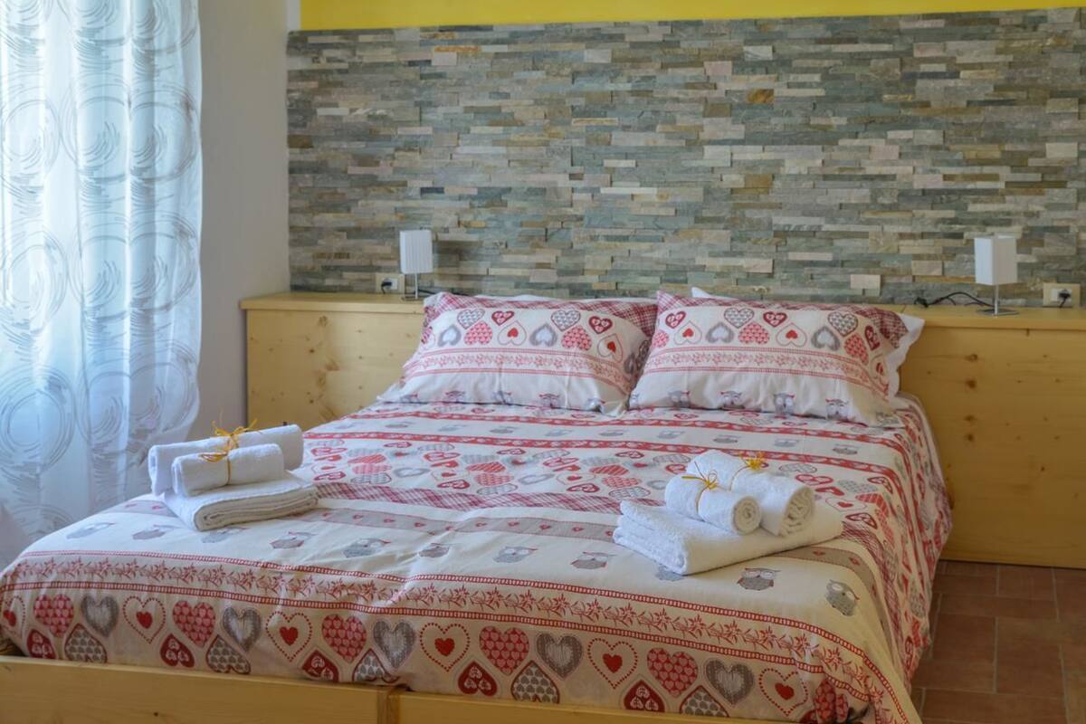 Una cama bien hecha con ropa de cama estampada y diseños de corazones, un cabecero de pared de piedra y una mesita de noche de madera con dos lámparas.