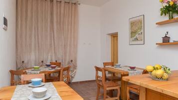 A dining area with wooden tables and chairs, a curtain, a wall-mounted picture, and a fruit basket.