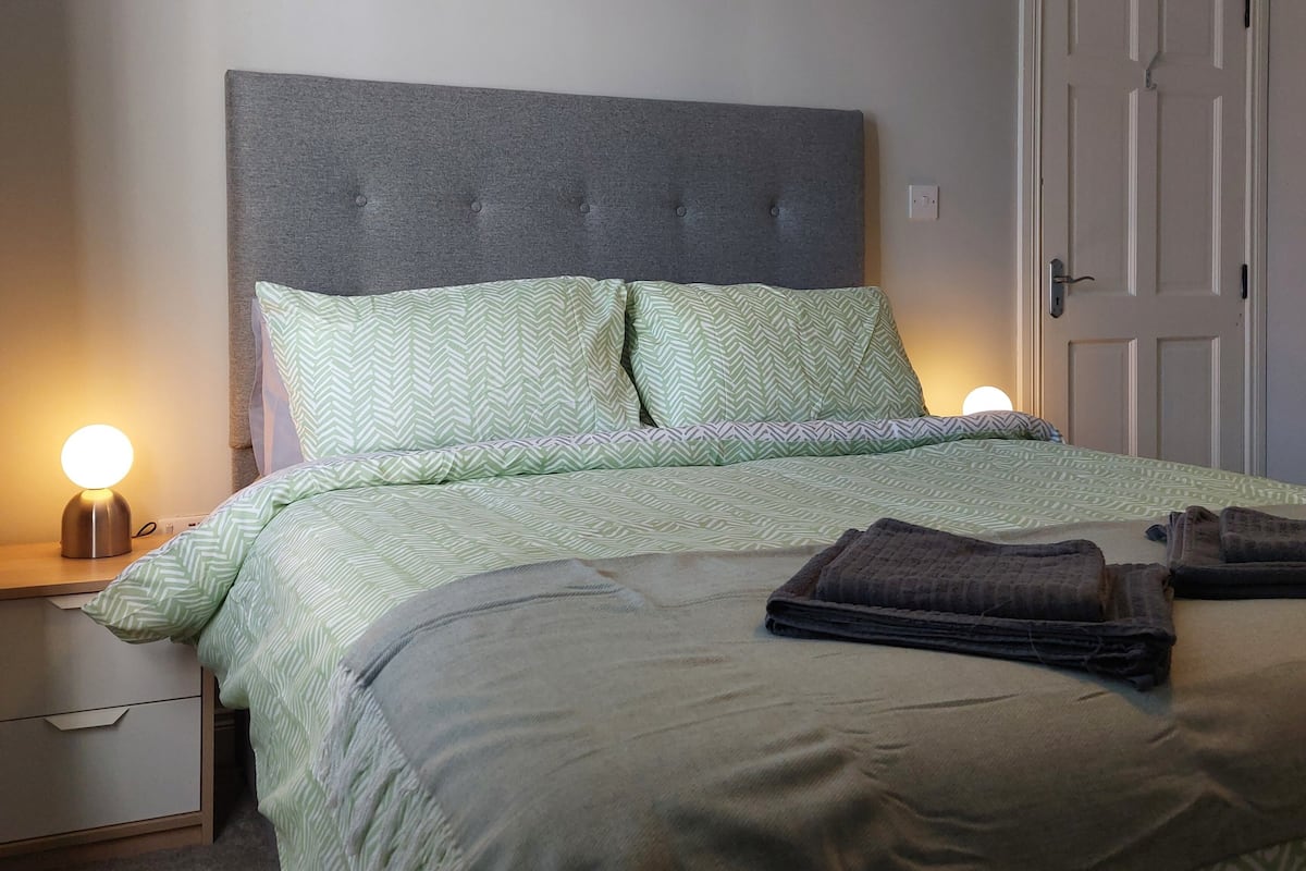 A neatly made bed with a grey headboard, green patterned bedding, and two bedside lamps.