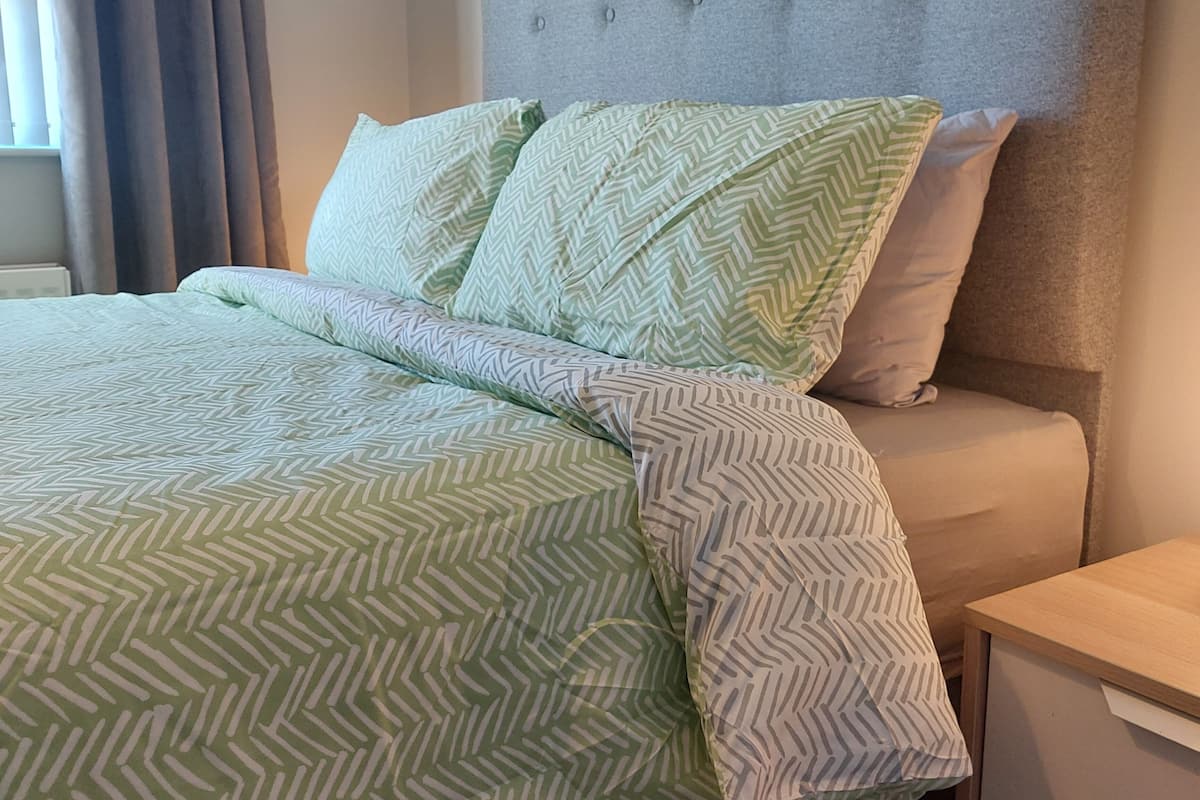 A neatly made bed with a green and white patterned bedspread and pillows, a grey upholstered headboard, and a wooden nightstand with a drawer.