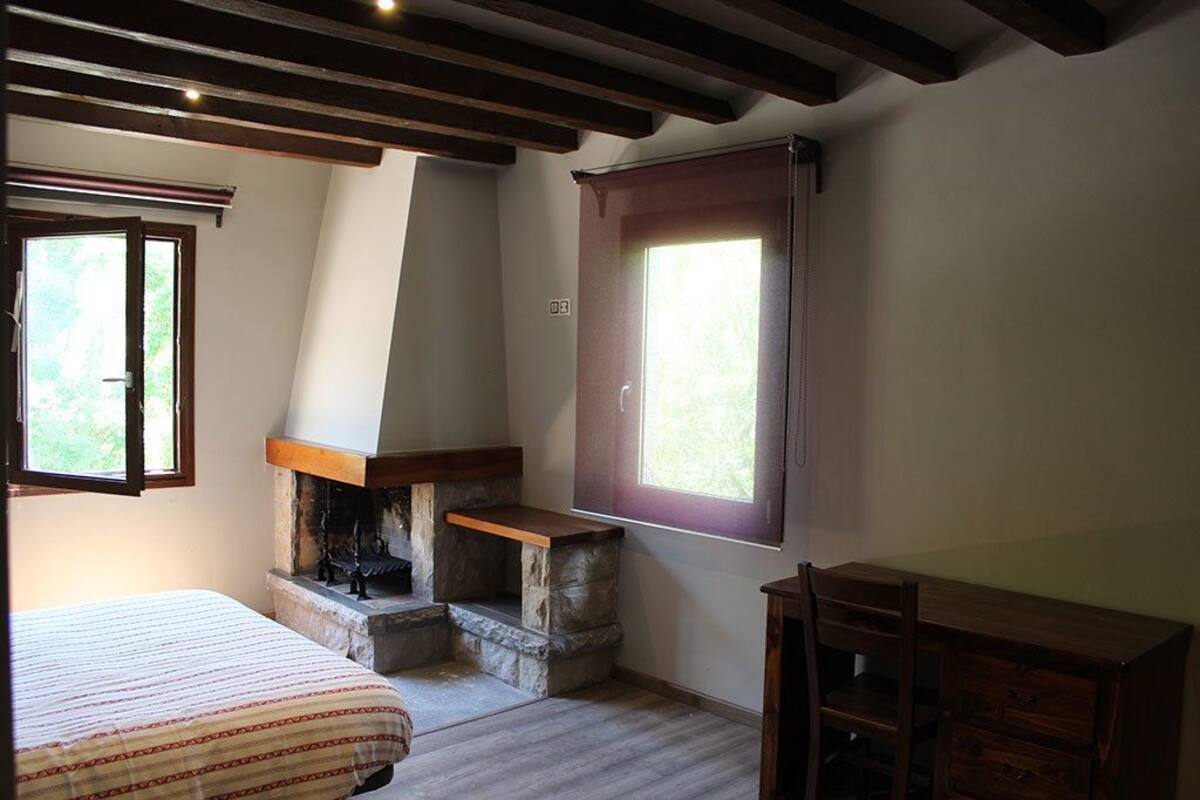 Un dormitorio con una cama, una chimenea de piedra, un aparador de madera y dos ventanas.