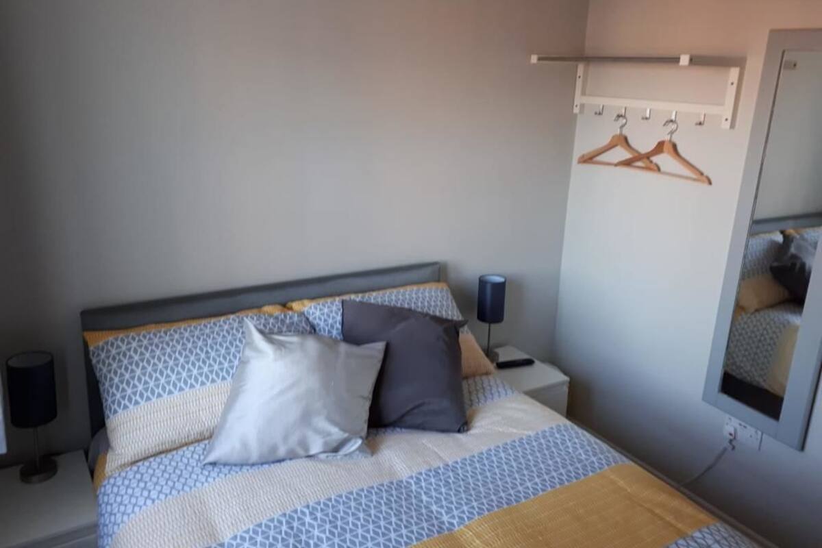 A neatly made bed with patterned bedding, a bedside table with a lamp, and a wall-mounted coat rack with hangers.