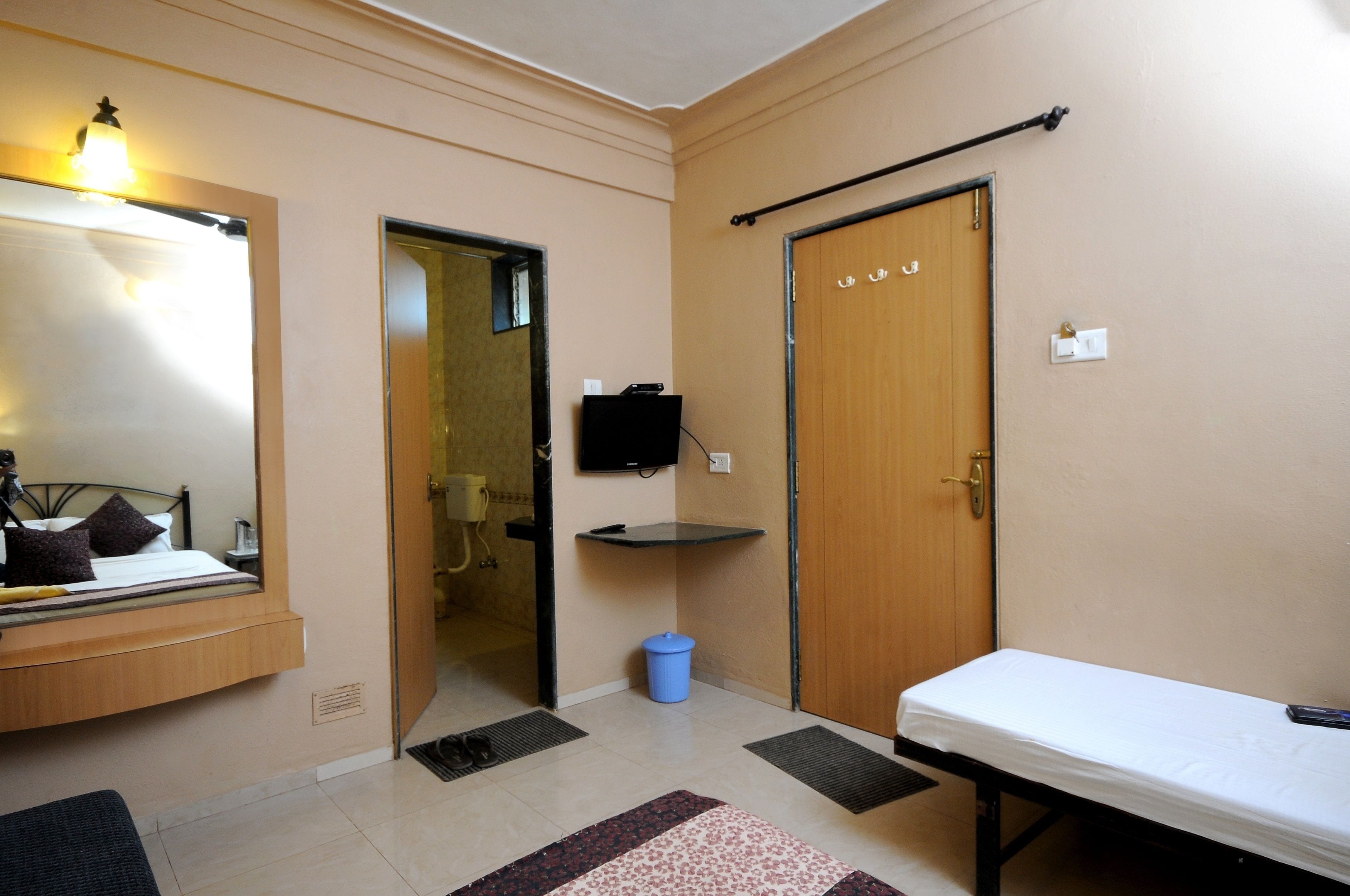 Une petite pièce bien éclairée, avec un lit, une table de chevet, un miroir, un téléviseur et une porte.