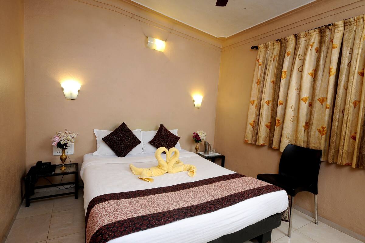 A hotel room with a neatly made bed, two bedside lamps, a nightstand with a phone, a chair, and a window with curtains.