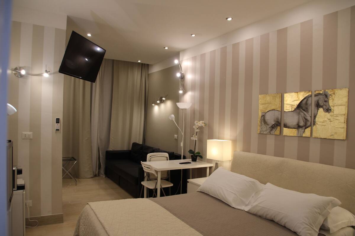 Ein Hotelzimmer mit Bett, Schreibtisch, Stuhl, einem an der Wand befestigten Fernseher und einem Gemälde von einem Pferd.