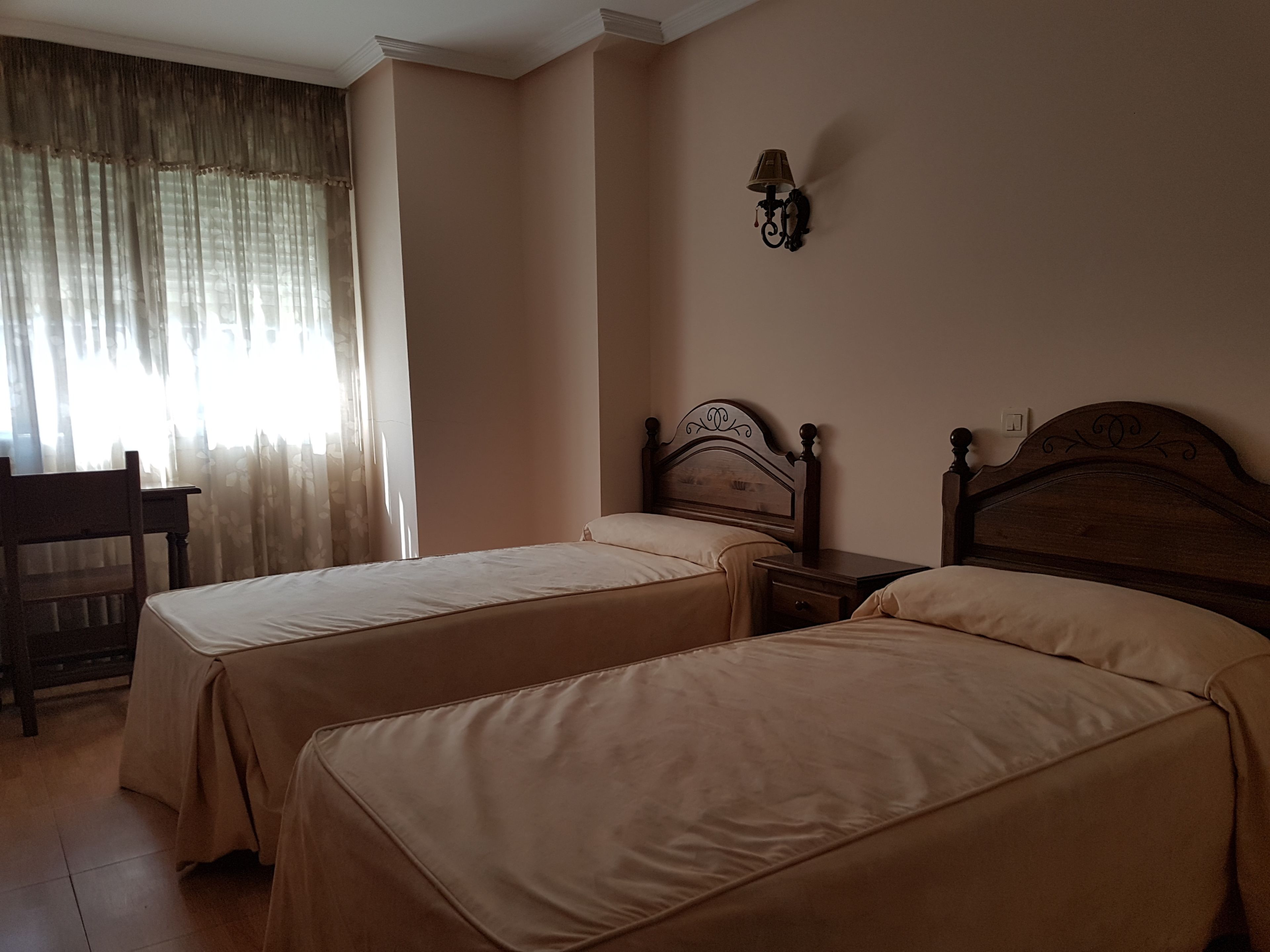 A bedroom with two single beds, a nightstand, a lamp, a chair, and a window with curtains.